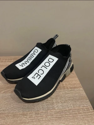 Dolce & Gabbana svarta slip-on sneakers - Svarta slip-on sneakers från Dolce & Gabbana med vit logga på ovansidan och detaljer i vitt och svart runt hälen. Ovandelen är i mesh-liknande material och sulan är svart med vita inslag. Snygg och sportig design som sticker ut. Kan gärna byta till ett annat par skor men bara om vi byter i person I Göteborg 