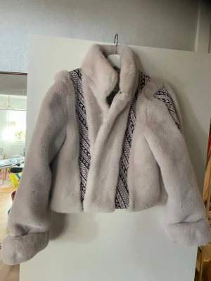 Vit pälsjacka med mönster - Säljer en fluffig vit  pälsjacka med bred krage och mönstrade band längs fram- och baksidan. Går att knäppa med små hakar. Jättefin!