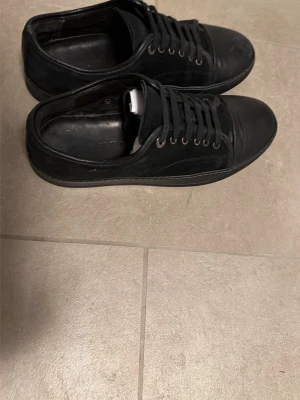 Svarta Lanvin sneakers  - Säljer ett par svarta lanvin sneakers med lågt skaft och klassisk design. Skorna har ovandel i mocka och svarta skosnören och metallfärgade öljetter. Perfekta för en clean och stilren look. Skicket är överlag bra. Lite små defekter. Se själv på bilderna! Hör av dig vid fråga😊