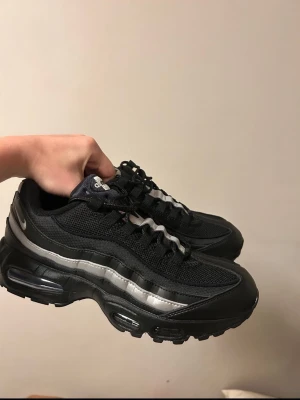 Nike Air Max 95 svarta sneakers HELT NYA - Nike Air Max 95 sneakers i svart med reflekterande silverdetaljer. Ovandel i mesh och syntet, synlig Air-enhet i sulan och klassisk vågig design. Swoosh-logga på hälen och tungans logotyp. Perfekta för dig som gillar streetwear och sportig stil.     Helt oanvända