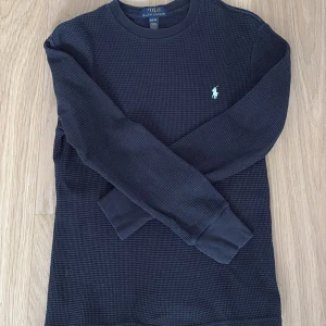 Svart långärmad topp Polo Ralph Lauren - Mörkblå långärmad topp från Polo Ralph Lauren med diskret rutstruktur i materialet och klassisk logga broderad på bröstet. Rund halsringning och mjuka muddar vid ärmslut. Perfekt för en clean och stilren look. Säljer den då den är för liten, skriv vid någon fråga!