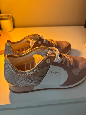 Rosa sneakers från Pepe Jeans - Snygga sneakers från Pepe Jeans i rosa och vitt med glittriga detaljer på hälen och sidan. Skorna har vita snören, vit sula och en klassisk låg modell. Perfekta för dig som vill ha en sportig och trendig look med lite extra bling.