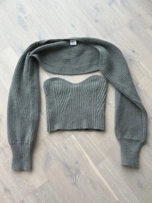 Grå stickad crop top med ärmar - Trendig grå stickad crop top med långa avtagbara ärmar. Ribbstickad design och snygg sweetheart-ringning. Tillverkad i 100% polyester, perfekt för att skapa en cool lager-på-lager-look.