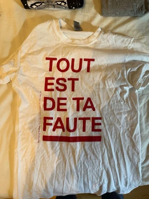 Vit t-shirt med röd text - Säljer en vit t-shirt med trycket 'TOUT EST DE TA FAUTE' i stora röda bokstäver på framsidan. T-shirten har rund hals och är i mjukt bomullsmaterial. Perfekt för dig som gillar statement-plagg och vill sticka ut med ett franskt budskap.