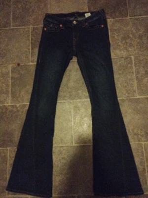 Mörkblå bootcut jeans med fickdetaljer - Säljer ett par mörkblå jeans med bootcut-modell och snygga fickdetaljer bak. Jeansen har kontrastsömmar och klassisk femficksdesign. Perfekta för dig som gillar en retroinspirerad look och vill ha ett par jeans som sticker ut lite extra.