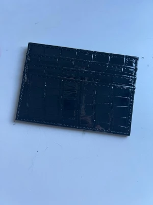 Svart kortfodral i krokopräglat skinn - Snyggt svart kortfodral med krokopräglat mönster och plats för flera kort. Tillverkat i glansigt konstläder som ger en lyxig känsla. Perfekt för dig som vill ha ett stilrent och smidigt sätt att bära dina kort.