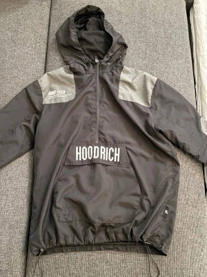 Hoodrich vindjacka - Snygg grå vindjacka från Hoodrich med halv dragkedja, stor huva och vita logotexter på bröstet och huvan. Jackan har ljusgrå paneler på axlarna, stor magficka och justerbar nederkant med dragsko. Tillverkad i lätt, vindtätt material. Skriv i dm för mer frågor 