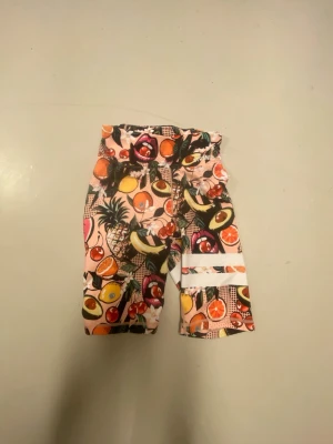 Färgglada träningsshorts med fruktmotiv - Säljer ett par färgglada träningsshorts med pop art-inspirerat fruktmotiv, bland annat avokado, apelsin, körsbär och ananas. Shortsen har vita ränder på ena benet och elastisk midja. Perfekta för gymmet eller löprundan.