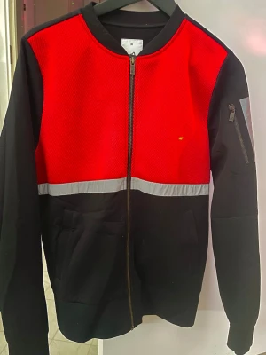 Röd och svart zip hoodie - Snygg kofta i storlek M. Framsidan är röd med svart nederdel och en grå rand som delar av färgerna. Dragkedja framtill, fickor och en extra ficka med dragkedja på ärmen. Tillverkad i mjukt material med meshstruktur upptill. Perfekt för en sportig look.