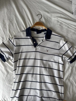 Vit och marinblå randig piké från Gant (M) - Snygg vit pikétröja från Gant med marinblå horisontella ränder, klassisk krage och knäppning framtill. Marinblå detaljer på krage och ärmslut samt liten Gant-logga broderad på bröstet. Knappt använd