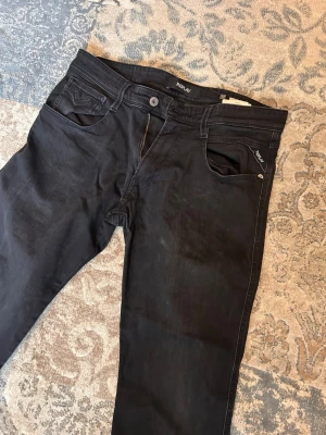 Svarta Replay jeans anbass - Säljer ett par svarta jeans från Replay, modell Anbass. Klassisk femficksdesign med dragkedjegylf och Replay-logga på fickan. Snygg passform och diskreta sömmar, perfekta till vardagslooken. Materialet är mjukt och stretchigt för extra komfort.