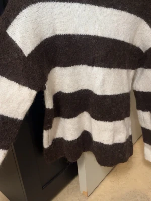 Randig fluffig stickad tröja - Mysig stickad tröja med breda bruna och vita ränder. Tröjan har en fluffig, mjuk yta och långa ärmar. Perfekt för dig som gillar en avslappnad och trendig look. 