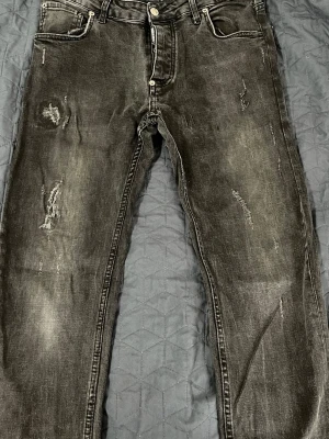 Svarta dsquared jeans med slitningar - Säljer ett par svarta jeans med coola slitningar och distressed detaljer. Jeansen har fem fickor, knappgylf och en något smal passform. Materialet är klassisk denim med en tvättad look som ger en trendig känsla. 