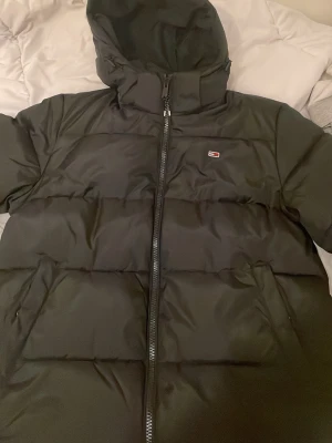 Svart pufferjacka från Tommy Hilfiger - Säljer en svart pufferjacka från Tommy Hilfiger med huva och dragkedja framtill. Jackan har ett litet Hilfiger-logomärke på bröstet och breda, vadderade paneler som ger en riktigt schysst look. Perfekt för kalla vinterdagar.