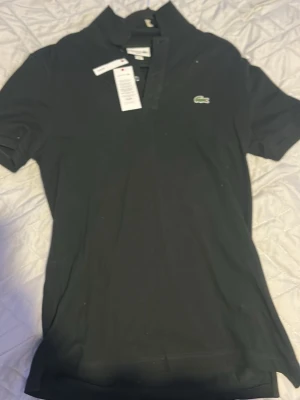 Svart pikétröja från Lacoste slim fit - Klassisk svart pikétröja från Lacoste i slim fit-modell. Tröjan har korta ärmar, krage och knappar framtill samt den ikoniska gröna krokodilloggan på bröstet. Tillverkad i mjuk bomull som känns skön mot huden. Helt oanvänd