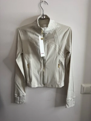 Vit zip-jacka från Lululemon storlek 4 - Snygg vit zip-jacka från Lululemon med guldfärgade dragkedjor och figurnära passform. Tillverkad i ett stretchigt funktionsmaterial som passar perfekt till träning eller som ett sportigt lager. Hög krage och två fickor framtill.