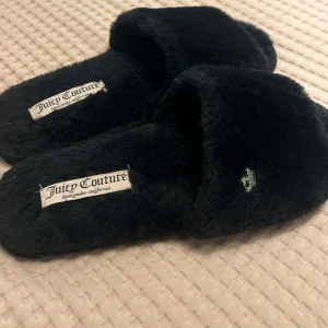 Svarta fluffiga Juicy Coutureh tofflor - Mysiga svarta tofflor från Juicy Couture i supermjukt, fluffigt material. Öppen tå och bred rem med liten metalllogga på sidan. Perfekta för att chilla hemma och ge en lyxig känsla till vardagen. Klassisk Juicy Couture-label på innersulan.