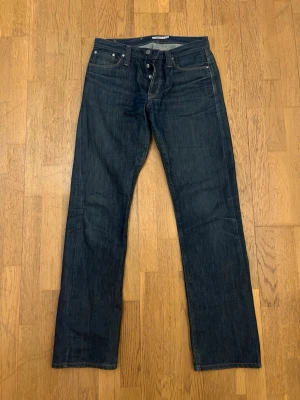 Mörkblå raka jeans från J.Lindeberg - strl.31 - Säljer ett par mörkblå jeans från J.Lindeberg med klassisk rak passform och fem fickor. Jeansen har kontrastsömmar och en patch bak i midjan. Tillverkade i kraftigt denimtyg som ger en tidlös look.