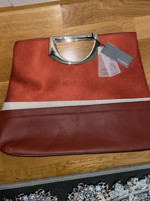 Rödbrun kuvertväska från Parfois - Snygg kuvertväska i rödbrunt med metallic handtag och en beige rand. Väskan har en mix av mockaimitation och slätt konstläder, vilket ger en cool kontrast. Perfekt accessoar för att lyfta din outfit med en modern touch.