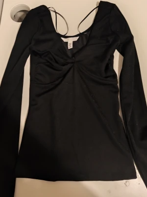Svart glittrig topp från H&M - Snygg svart långärmad topp från H&M med glittriga detaljer och djup v-ringning. Toppen har en twistad detalj framtill och tunna axelband som ger en unik look. Perfekt för dig som vill sticka ut lite extra.