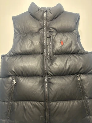 Svart dunväst från Polo Ralph Lauren - Snygg svart dunväst från Polo Ralph Lauren med hög krage och tre dragkedjefickor. Västen har ett diskret rött logobroderi på bröstet och quiltad design. Perfekt för lager-på-lager och streetwear-vibe. Materialet är glansigt och ger en modern look. Västen är köpt på kidsbrande store för 2800kr. Vid minsta frågor är det bara att skriva. Priser är ej fast.