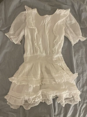Vit volangklänning med puffärm - Supersöt vit kortklänning med volanger och puffärmar. Klänningen har markerad midja med smock och flera lager volanger nedtill.❤️