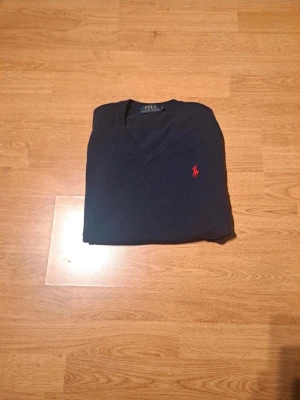 BUD stickad tröja Polo Ralph Lauren  - Mörkblå stickad tröja från Polo Ralph Lauren med klassisk V-ringning och röd broderad logga på bröstet. Tröjan har långa ärmar och ribbade muddar vid ärmslut och nederkant. Perfekt för lager på lager och stilren look.