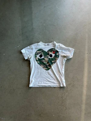 Comme des Garçons t-shirt - Vit t-shirt från Comme des Garçons med ett stort hjärta i camouflage-mönster och ögon på bröstet. Liten röd logga med hjärta på vänster sida. Klassisk passform och korta ärmar. Perfekt statement-plagg för dig som gillar streetwear. Skriv vid frågor