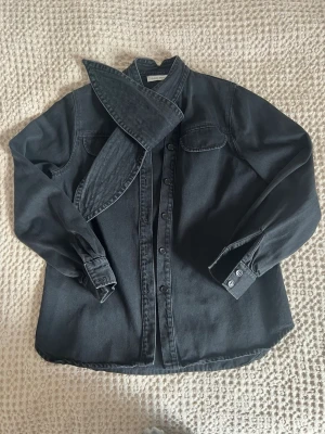 Svart jeansjacka med stor krage - Säljer en svart jeansjacka från med knappstängning framtill. Jackan har två bröstfickor med lock och långa ärmar med knappar vid ärmslut. Snygg och trendig modell som sticker ut med sin unika krage.
