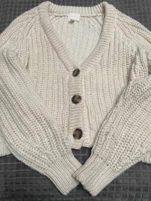 Beige stickad kofta med knappar - Säljer en chunky beige stickad kofta med stora bruna knappar framtill. Koftan har v-ringning, bred ribbstickad kant och långa, lite pösiga ärmar. Perfekt för lager på lager och riktigt mysig att ha på sig.