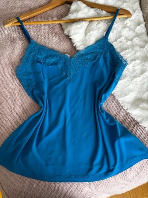 Blått linne - Snygg blå linnetopp med tunna axelband och v-ringad spetskant framtill. Materialet känns lent mot huden och spetsen ger en extra touch. Storlek XS men känns mer som S och M