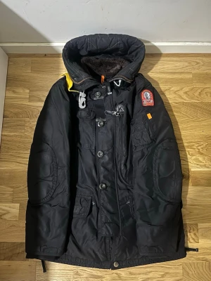 Svart Parajumpers parkajacka  - Svart parkajacka från Parajumpers med stor huva, gul detalj på axeln och flera fickor. Jackan har både dragkedja och knappar framtill, quiltad insida och mjuk fuskpäls vid kragen. Perfekt för kalla dagar och har coola detaljer som patch på ärmen.helt äkta 
