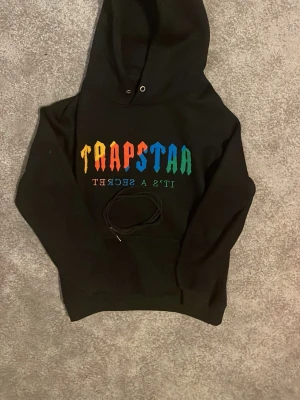 Trapstar svart hoodie med färgglad text - Svart hoodie från Trapstar med stor huva och magficka. Framsidan har färgglad text i regnbågens färger där det står 'TRAPSTAR' och 'IT'S A SECRET' spegelvänt under. Tillverkad i mjukt material, perfekt för en chill och streetwear-inspirerad look. Priset kan sänkas.