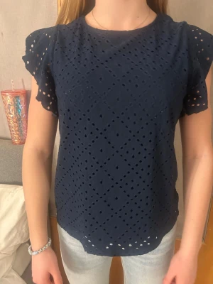 Mörkblå blus med broderade hål - Snygg mörkblå blus med korta volangärmar och broderade hål i ett diamantmönster. Tillverkad i bomull som känns lätt och luftig. Perfekt till jeans eller kjol.