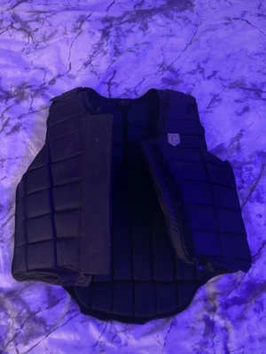 Svart quiltad skyddsväst för ridning - Svart quiltad skyddsväst med rutmönster och justerbar passform. Västen har en diskret logga på bröstet och stängs med kardborre framtill. Insidan har en röd detalj. Perfekt för dig som vill ha extra skydd vid ridning.