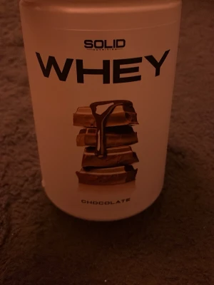 Solid Nutrition Whey Chocolate - Säljer en burk Solid Nutrition Whey proteinpulver med chokladsmak. Förpackningen är vit med svart text och har en bild på chokladbitar. Innehåller 750g och är laktosfri. Perfekt för dig som tränar och vill ha extra protein. Det är mer än halva kvar