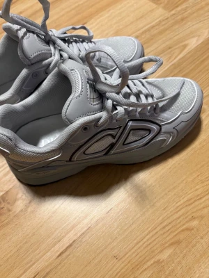 Grå Dior sneakers med CD-detalj - Snygga grå sneakers från Dior med stora CD-detaljer på sidan och klassisk snörning. Skorna har en chunky sula och är tillverkade i en mix av mesh och syntetmaterial för en modern look. Perfekta för dig som vill ha en exklusiv touch till din outfit.