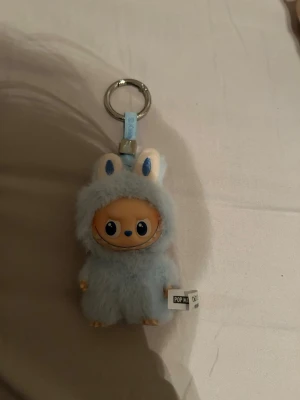 Pop Mart blå fluffig nyckelring - Söt Labubu nyckelring från Pop Mart med en figur i blå fluffig dräkt med kaninöron. Ansiktet är i plast och dräkten är mjuk och lurvig. Perfekt att hänga på väskan eller nycklarna för en gullig touch.