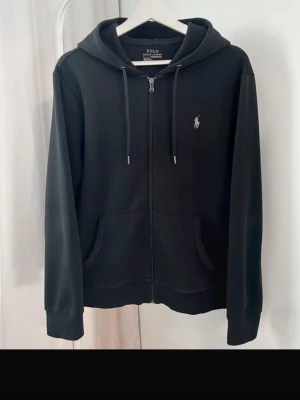 Svart zip hoodie från Polo Ralph Lauren - Säljer en svart hoodie med dragkedja från Polo Ralph Lauren. Klassisk design med huva, fickor framtill och den ikoniska vita logon broderad på bröstet. Tillverkad i mjukt bomullsmaterial som är skönt att bära. Perfekt för en avslappnad och stilren look.