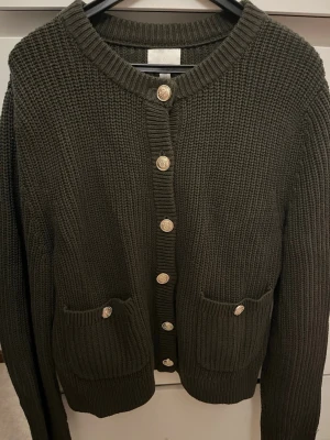 Mörkgrön stickad kofta från H&M - Snygg mörkgrön stickad kofta från H&M med guldfärgade knappar framtill och två fickor med knappdetaljer. Koftan har rund halsringning och ribbade muddar vid ärmslut och nederkant. Perfekt för lager på lager och en chill look.