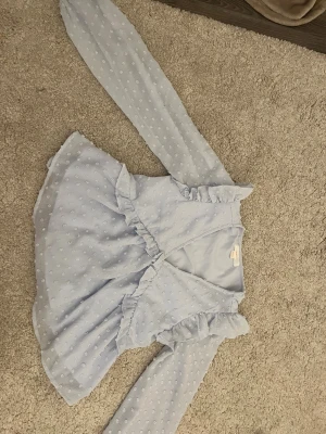 Ljusblå volangblus från Gina Tricot - Supersöt ljusblå blus från Gina Tricot med volangdetaljer och diskret prickmönster. Blusen har omlott-look framtill och långa ärmar. Perfekt för dig som gillar romantisk och trendig stil. Materialet känns lätt och luftigt.