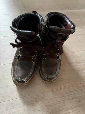 Bruna vinter kängor - Snygga bruna kängor med högt skaft och grov sula. Kängorna har vita kontrastsömmar, metallhakar för snörning och vadderad kant upptill. Perfekta för höst och vinter, tillverkade i läder med robust känsla.