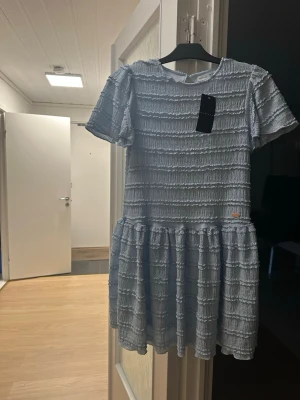 Ljusblå kortklänning från Sumwon - Supersöt ljusblå kortklänning från Sumwon med strukturmönster och volangdetaljer. Klänningen har rund hals, korta ärmar och en avslappnad passform. Perfekt för dig som gillar en lekfull och trendig stil.