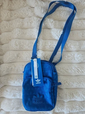 Blå festivalväska från Adidas - Snygg blå axelväska från Adidas med justerbar rem och ikoniskt Trefoil-märke framtill. Helt ny med lappen kvar