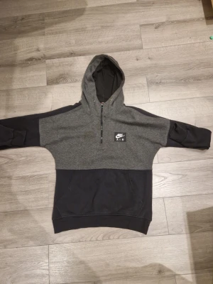 Nike Air tvåfärgad hoodie S - Snygg tvåfärgad hoodie från Nike Air i mörkgrått och svart. Tröjan har halv dragkedja framtill, stor huva och en praktisk magficka. Tillverkad i mjukt bomullsmaterial och har Nike Air-logga på bröstet. Perfekt för en avslappnad streetwear-look.