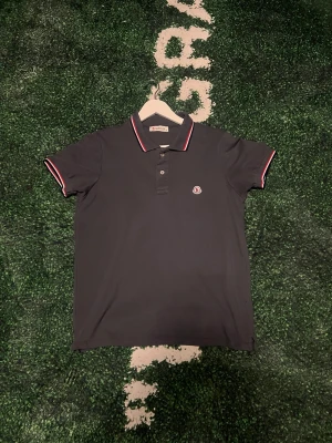 Svart pikétröja från M - Klassisk svart pikétröja från M med korta ärmar och knappslå framtill. Kragen och ärmsluten har vita och röda ränder för en sportig touch. M-logga på bröstet. Perfekt till en avslappnad men stilren look.