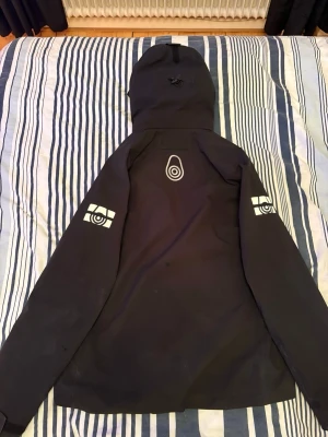 Svart vindjacka från Peak Performance - Svart vindjacka med huva från sail racing. Jackan har vita grafiska tryck på ryggen och ärmarna, meshfoder på insidan och dragkedja framtill. Perfekt för blåsiga dagar och har en modern, sportig look.