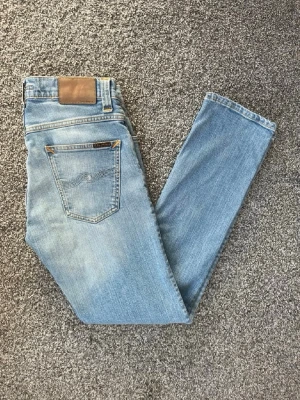 Ljusblå  jeans från Nudie - Säljer ett par ljusblå sjeans från Nudie Jeans med klassisk femficksmodell och läderpatch bak i midjan. Jeansen har en snygg tvätt och subtila slitningar för en avslappnad look. Tillverkade i bomull med stretch för extra komfort.