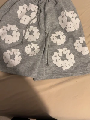 Grå shorts med vita tryck och snörning - Snygga grå shorts med elastisk midja och snörning. Shortsen har flera vita grafiska tryck över hela plagget och är perfekta för en avslappnad stil. Tillverkade i mjukt material som känns skönt mot huden.