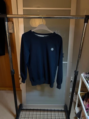 Mörkblå sweatshirt från Stone Island - Snygg mörkblå sweatshirt från Stone Island med klassisk rund halsringning och ribbade muddar. Tröjan har den ikoniska Stone Island-loggan på vänstra ärmen och en liten patch på bröstet. Perfekt för en stilren och avslappnad look.